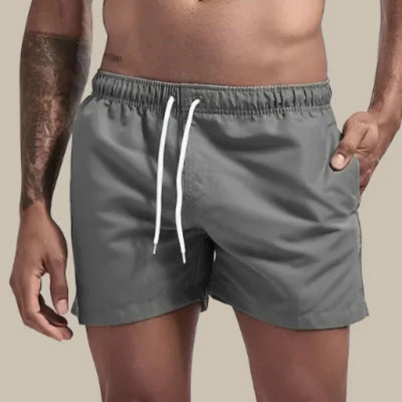 Laden Sie das Bild in Galerie -Viewer, Mann in grauer Badehose mit Kordelzug, tätowierter Arm, neutraler Hintergrund. Herrenmode, Sommerbekleidung, Strandmode.
