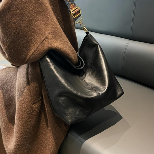 Schwarze Ledertasche auf grauem Sofa, brauner Mantel im Hintergrund. Modische Handtasche, elegantes Design, hochwertiges Leder, Damenaccessoire.