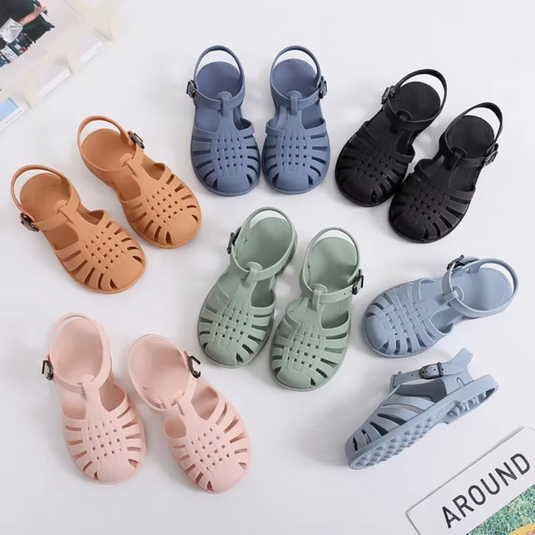 Mehrfarbige Kinder-Sandalen aus Kunststoff, in Blau, Schwarz, Beige, Rosa und Grün, auf weißem Hintergrund angeordnet. Ideal für Sommermode.
