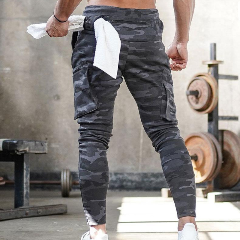 Laden Sie das Bild in Galerie -Viewer, Mann in Camouflage-Trainingshose im Fitnessstudio, hält ein Handtuch. Hantelbank und Gewichte im Hintergrund. Sportbekleidung, Fitness, Workout.
