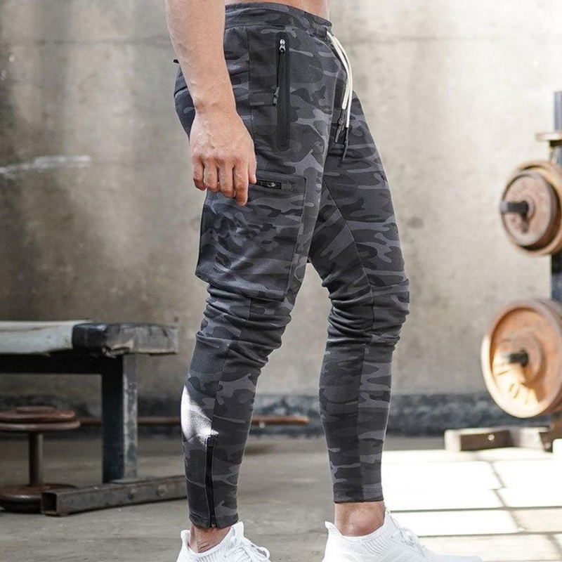 Laden Sie das Bild in Galerie -Viewer, Mann in grauer Camouflage-Jogginghose im Fitnessstudio, Hanteln im Hintergrund. Sportbekleidung, Fitnessmode, Herren-Trainingshose.
