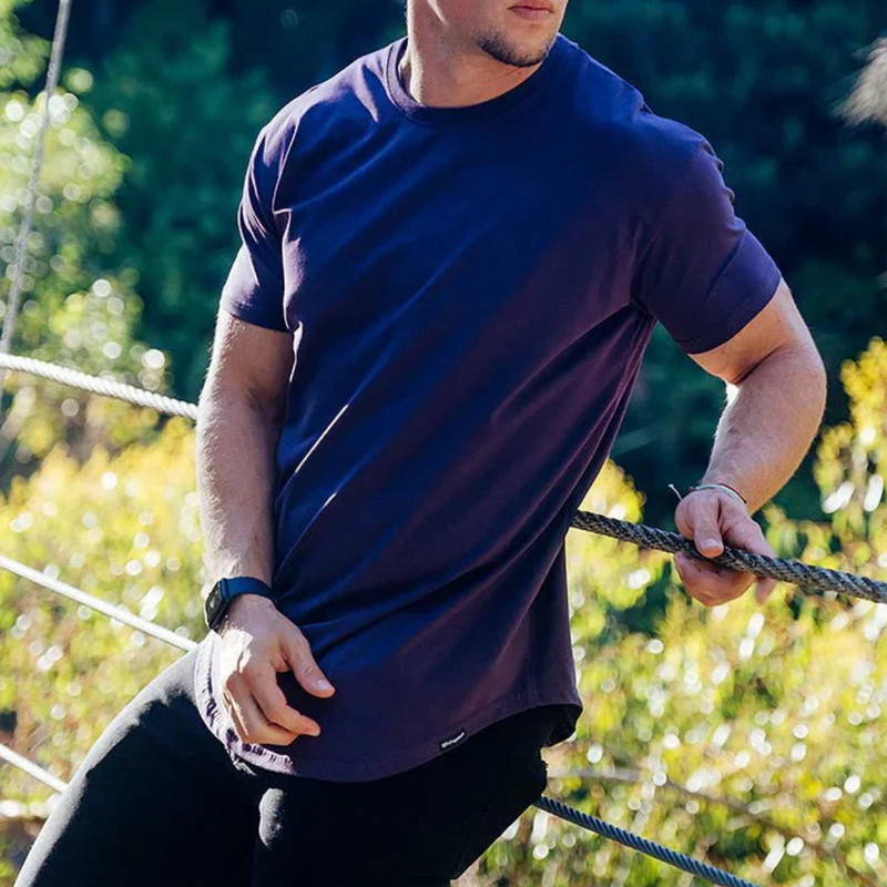 Laden Sie das Bild in Galerie -Viewer, Ein Mann in einem dunkelblauen T-Shirt lehnt an einem Seil im Freien. Er trägt eine schwarze Uhr. Hintergrund mit grüner Natur.
