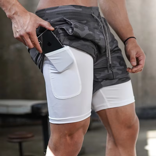 Mann in grauen Camouflage-Trainingsshorts mit integriertem Handyfach, hält Smartphone. Sportbekleidung, Fitnessmode, praktische Sportshorts.