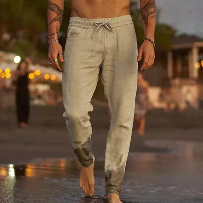 Mann in nasser, hellgrauer Jogginghose am Strand bei Sonnenuntergang, barfuß im Wasser. Freizeitmode, Strandspaziergang, Sommerabend.