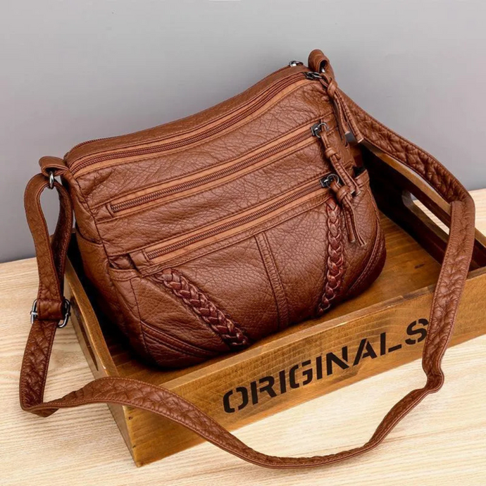 Braune Lederhandtasche mit mehreren Reißverschlusstaschen und geflochtenen Details, auf Holzkiste liegend. Modische Damen-Umhängetasche.
