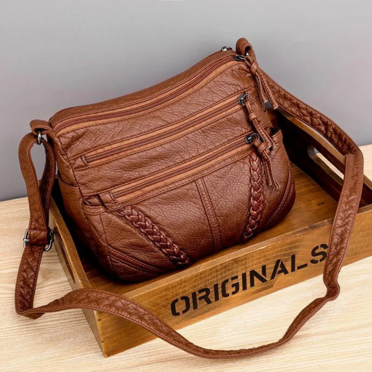 Braune Lederhandtasche mit mehreren Reißverschlusstaschen und geflochtenen Details, auf Holzkiste liegend. Modische Damen-Umhängetasche.