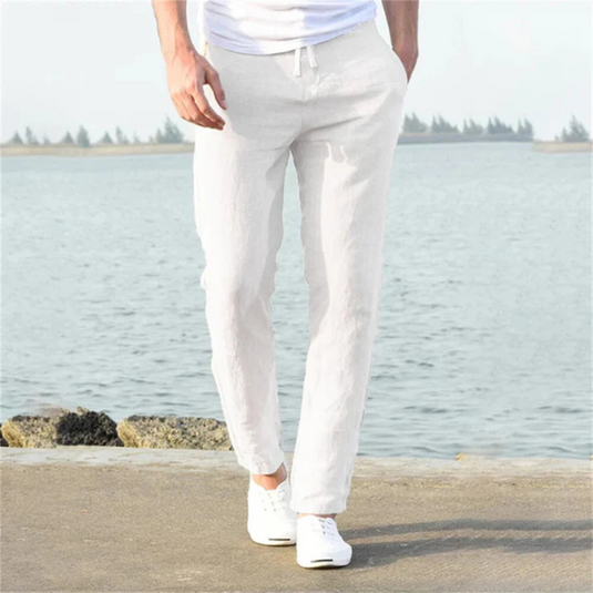 Mann in weißen Leinenhosen und weißen Sneakers steht am Wasser. Sommermode, Freizeitlook, Herrenbekleidung, stilvoll, entspannt.