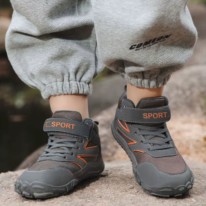 Laden Sie das Bild in Galerie -Viewer, Graue Kindersportschuhe mit Klettverschluss und orangefarbenen Akzenten, getragen von einem Kind in grauer Jogginghose. Outdoor-Abenteuer, bequeme Kinderschuhe.
