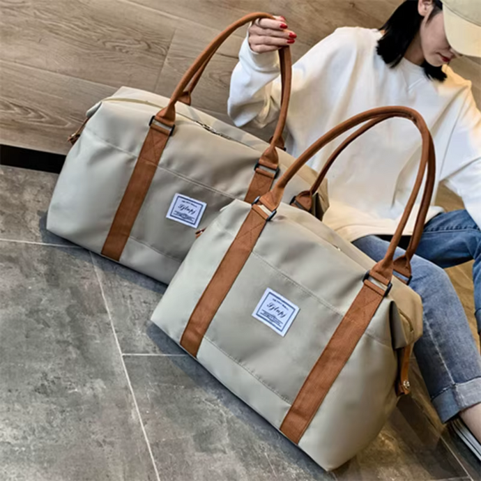 Zwei beige Reisetaschen mit braunen Griffen auf grauem Boden, Frau im Hintergrund. Stilvolle, geräumige Taschen für Reisen und Alltag.