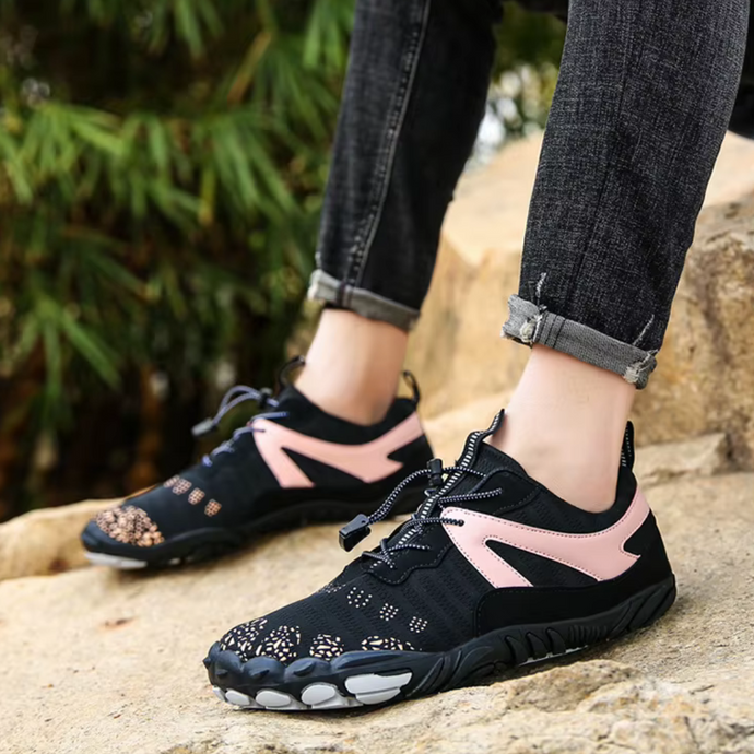 Schwarze Barfußschuhe mit rosa Akzenten auf Felsen, getragen von Person in hochgekrempelten Jeans. Outdoor-Schuhe, minimalistisch, atmungsaktiv.