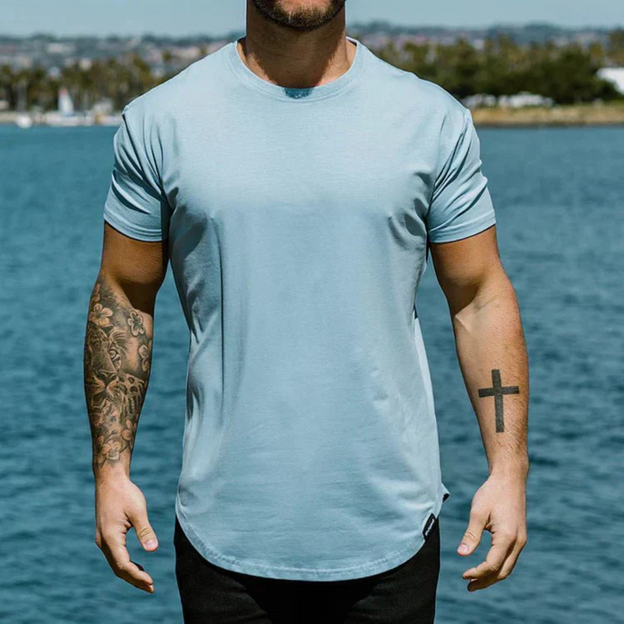 Mann in hellblauem T-Shirt mit Tätowierungen steht vor Wasserlandschaft. Sommermode, Herrenbekleidung, Freizeitlook, Tätowierungen, See im Hintergrund.