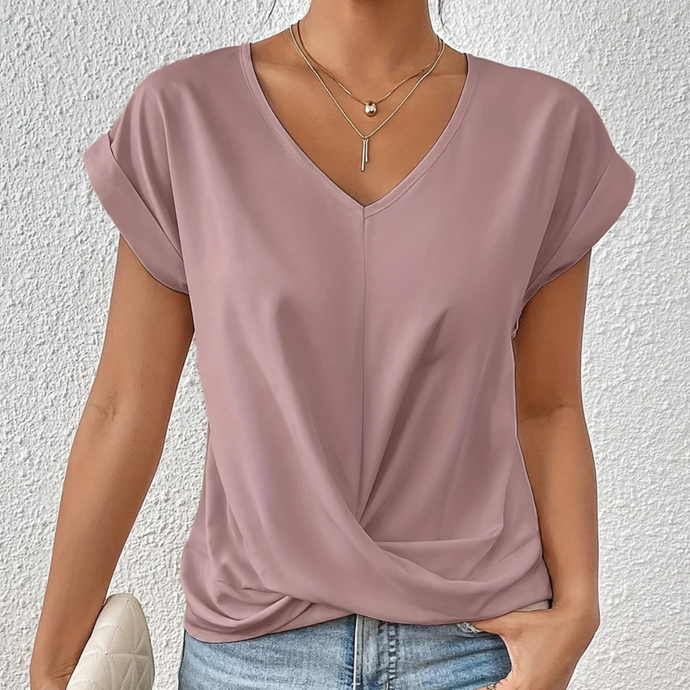 Frau trägt lila V-Ausschnitt T-Shirt, kombiniert mit blauen Jeans und goldener Halskette, vor weißer Wand. Modetrend, lässiger Stil.