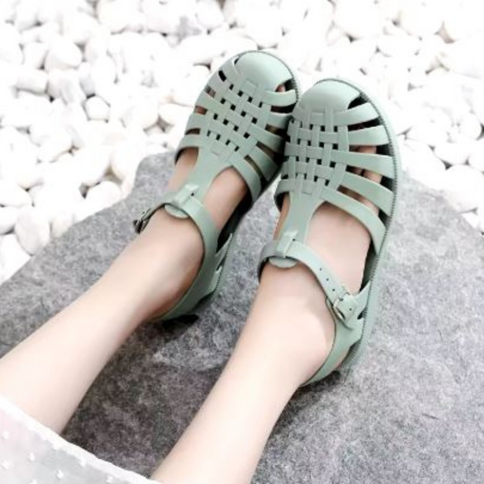 Damenfüße in mintgrünen, geflochtenen Sandalen auf grauem Stein, umgeben von weißen Kieselsteinen. Sommermode, bequeme Schuhe, Outdoor-Stil.