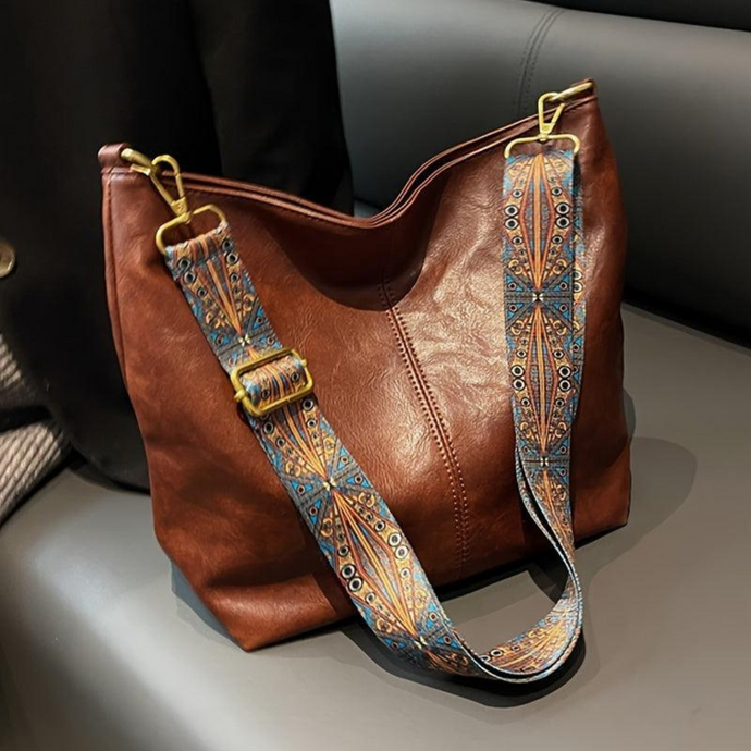 Braune Ledertasche mit verziertem, buntem Schultergurt auf grauem Sofa. Modische Handtasche, elegantes Design, hochwertiges Accessoire.