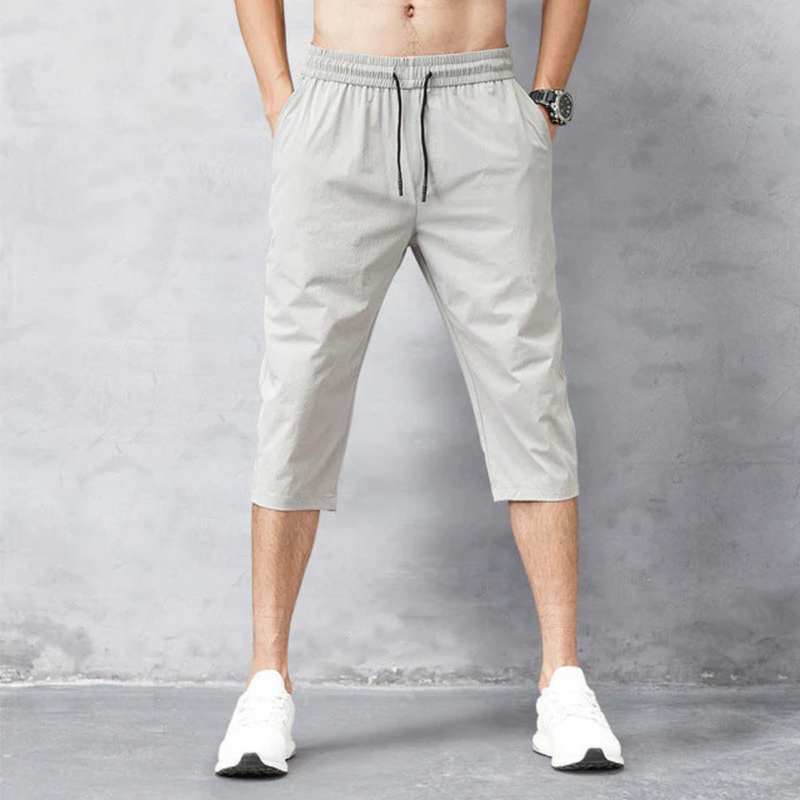 Laden Sie das Bild in Galerie -Viewer, Mann trägt graue, knielange Jogginghose und weiße Sneaker vor grauem Hintergrund. Sportliche Herrenmode, bequeme Freizeitkleidung.
