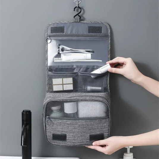 Graue hängende Kulturtasche mit mehreren Fächern, gefüllt mit Reiseutensilien wie Zahnbürste, Rasierer und Kosmetik, an grauer Badezimmerwand.