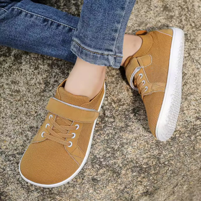 Braune Sneaker mit Klettverschluss und Schnürsenkeln, getragen von Person in blauen Jeans, auf grauem Stein. Bequeme Freizeitschuhe für Damen.