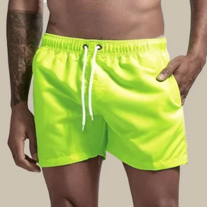 Mann in neongrüner Badeshorts mit Kordelzug, tätowierter Arm, neutraler Hintergrund. Sommermode, Herrenbadehose, Strandbekleidung.