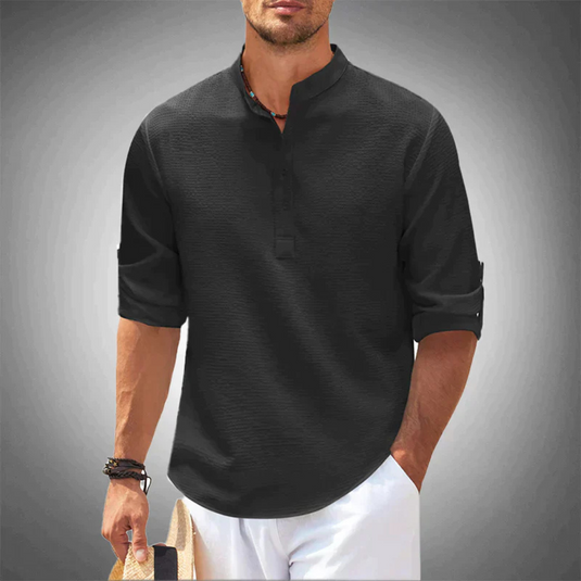 Mann in schwarzem Henley-Shirt mit hochgekrempelten Ärmeln, hält Strohhut, vor grauem Hintergrund. Herrenmode, Freizeitlook, stilvoll.