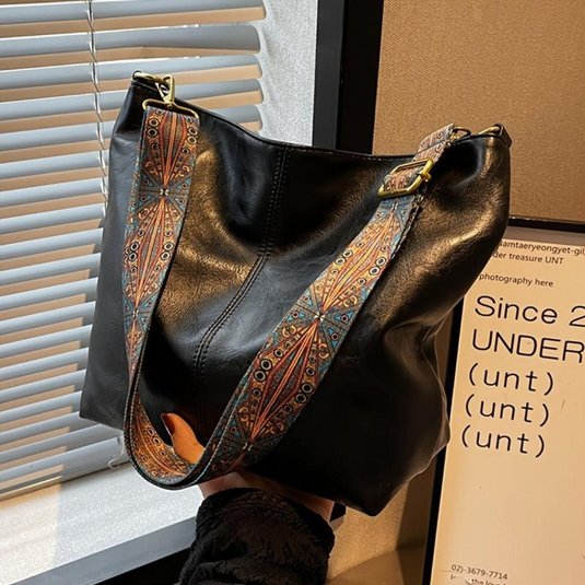 Schwarze Ledertasche mit buntem, gemustertem Schulterriemen, vor einem Fenster mit Jalousien. Elegante Handtasche, modisches Accessoire.