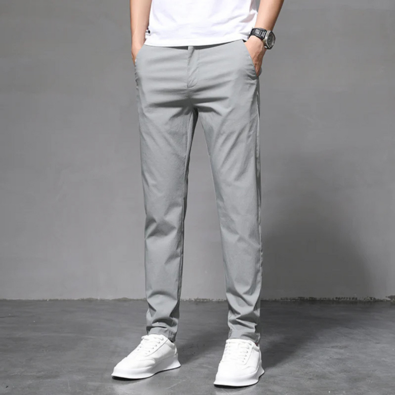 Laden Sie das Bild in Galerie -Viewer, Mann in grauer Chinohose und weißen Sneakers vor grauem Hintergrund, lässiger Stil, moderne Herrenmode, Freizeitlook, stilvolle Kleidung.
