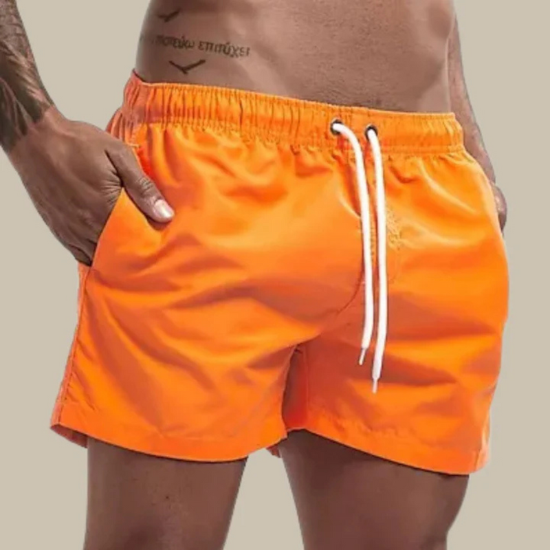 Laden Sie das Bild in Galerie -Viewer, Mann in leuchtend orangefarbener Badehose mit Kordelzug, Hand in der Tasche, vor neutralem Hintergrund. Sommermode, Herrenbadebekleidung.
