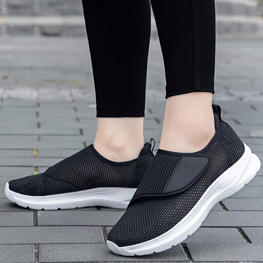 Schwarze Slip-On-Sneaker mit weißer Sohle auf grauem Pflaster, getragen von Person in schwarzer Leggings. Bequeme, atmungsaktive Freizeitschuhe.