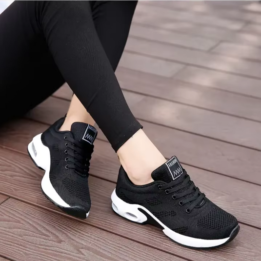 Schwarze Damen-Sneaker mit weißer Sohle, atmungsaktives Mesh-Design, getragen von Person in schwarzer Leggings auf Holzterrasse.
