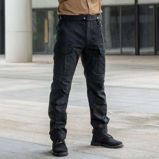 Mann in schwarzer taktischer Hose und Stiefeln steht draußen. Outdoor-Bekleidung, robuste Cargohose, funktionale Mode für Abenteuer.