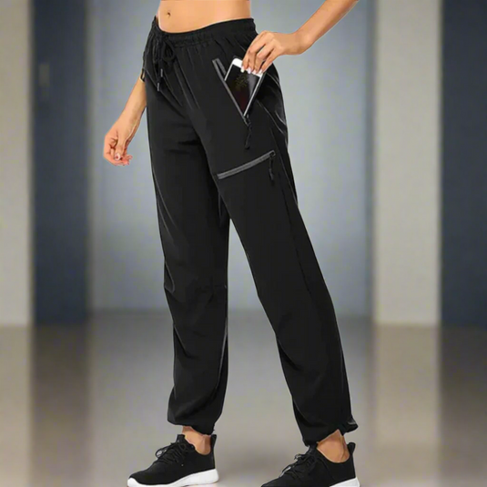 Frau in schwarzer Sporthose mit Smartphone in der Tasche, steht in modernem Raum. Fitnessmode, bequeme Jogginghose, praktisches Design.