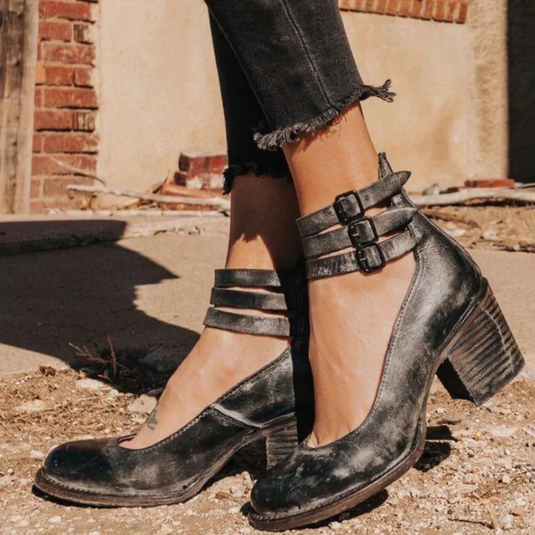 Schwarze Vintage-Lederstiefeletten mit Blockabsatz und Schnallen, getragen von einer Person in abgeschnittenen Jeans, auf urbanem Hintergrund.