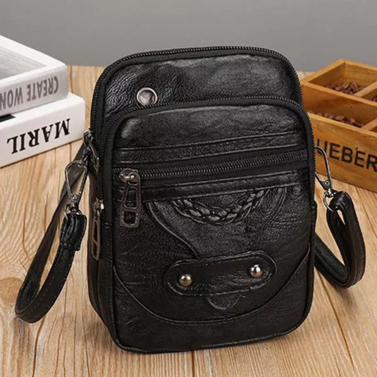 Schwarze Leder-Umhängetasche auf Holztisch, mit Reißverschlüssen und Nieten, ideal für Herren, kompakt und stilvoll, perfekt für Alltag und Reisen.