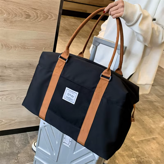 Dunkle Reisetasche mit braunen Griffen auf einem Koffer, gehalten von einer Person in weißem Pullover. Perfekt für Reisen und Wochenendausflüge.