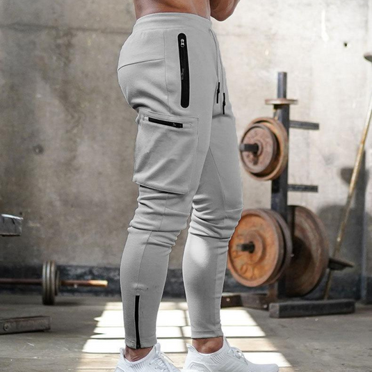 Graue Jogginghose mit Taschen, Reißverschlüssen, im Fitnessstudio vor Hanteln. Sportbekleidung, Herrenmode, funktionale Trainingshose.