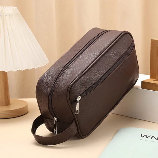Braune Leder-Kulturtasche mit Reißverschluss, auf Tisch neben Lampe und Buch. Praktisch für Reisen, stilvolles Design, Herren-Accessoire.