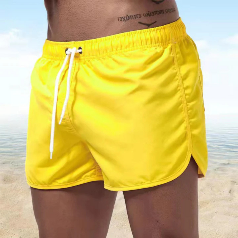 Laden Sie das Bild in Galerie -Viewer, Mann in gelber Badehose am Strand, blauer Himmel im Hintergrund. Sommermode, Strandbekleidung, Herrenbadehose, Urlaubsoutfit.

