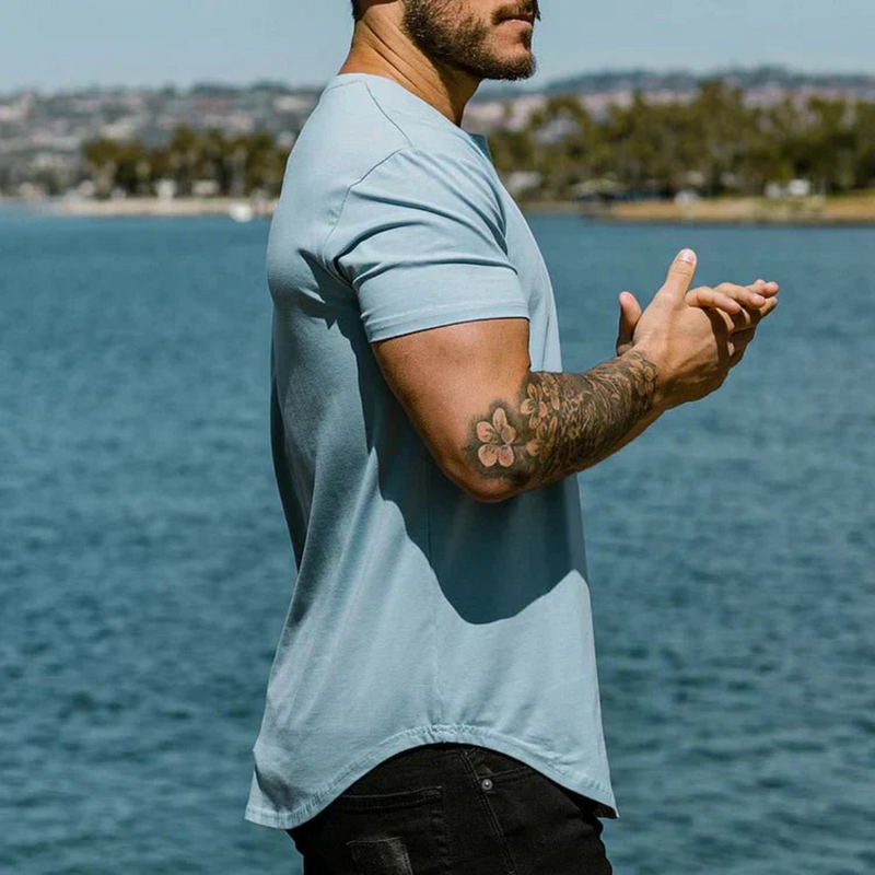 Laden Sie das Bild in Galerie -Viewer, Mann im blauen T-Shirt mit tätowiertem Arm steht am Seeufer, sommerliche Landschaft im Hintergrund. Freizeitmode, Tätowierungen, Natur.
