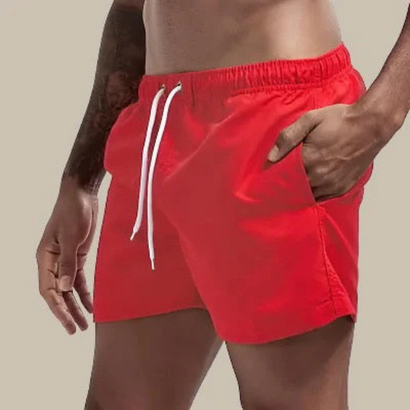 Laden Sie das Bild in Galerie -Viewer, Mann in roten Badeshorts mit Kordelzug, Hand in der Tasche, tätowierter Arm, sportlicher Körperbau. Ideal für Strandmode und Sommerbekleidung.
