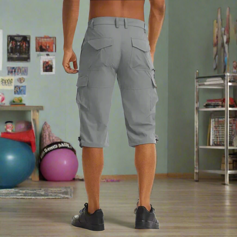 Laden Sie das Bild in Galerie -Viewer, Mann in grauen Cargoshorts von hinten in einem Fitnessraum mit Gymnastikbällen und Regalen, sportliche Kleidung, Freizeitmode, Fitnessstudio.
