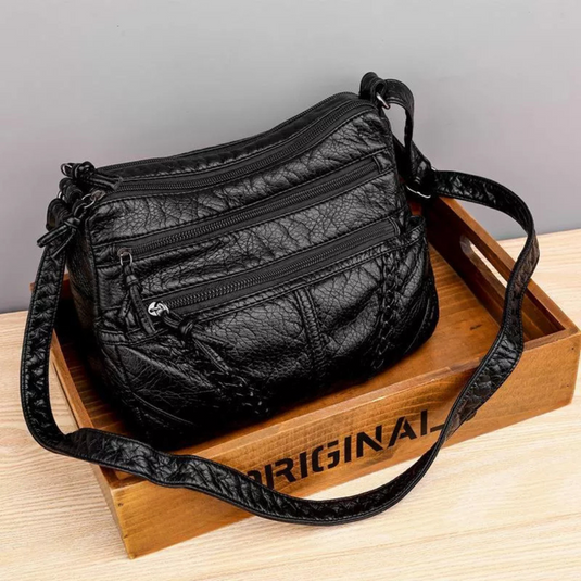 Schwarze Lederhandtasche mit mehreren Reißverschlusstaschen auf Holzkiste. Modische Damenhandtasche, ideal für Alltag und Reisen.