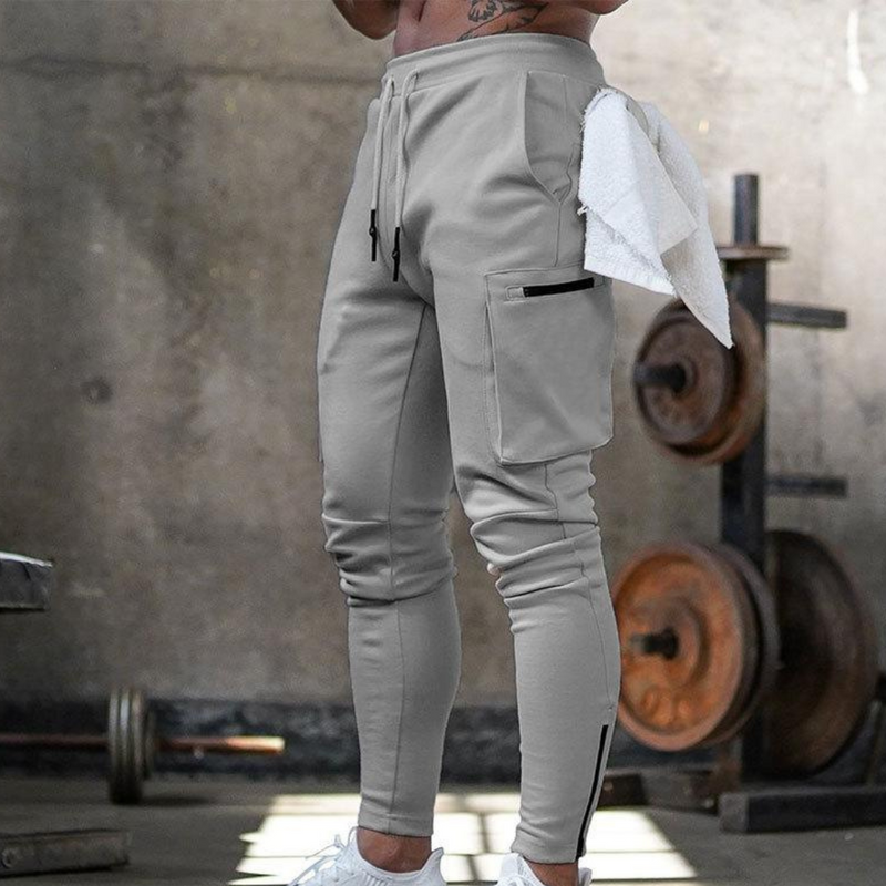 Laden Sie das Bild in Galerie -Viewer, Graue Jogginghose mit Taschen, getragen von einem muskulösen Mann im Fitnessstudio. Hantel im Hintergrund. Sportbekleidung, Fitness, Training.
