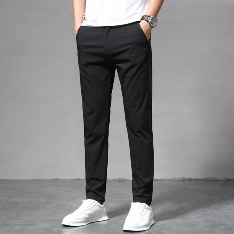 Laden Sie das Bild in Galerie -Viewer, Mann in schwarzer Chinohose und weißen Sneakers vor grauem Hintergrund. Modische Herrenbekleidung, lässiger Stil, moderne Herrenhose.
