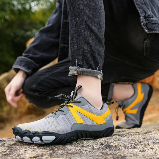 Graue und gelbe Barfußschuhe auf Felsen, getragen von Person in schwarzer Jeans. Outdoor-Schuhe, minimalistisch, flexibel, ideal für Naturwanderungen.