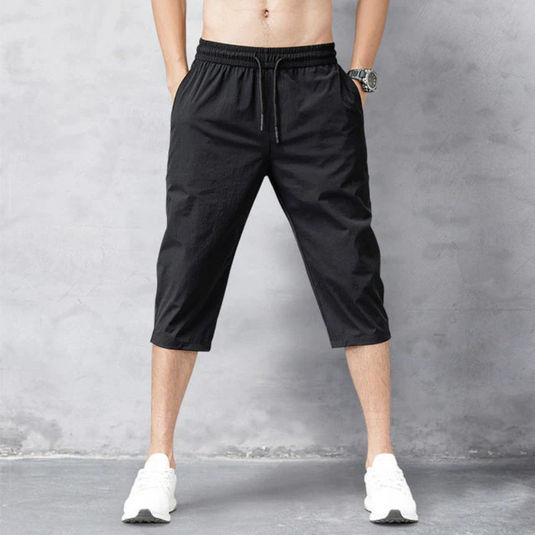 Mann in schwarzen Dreiviertelhosen und weißen Sneakers vor grauem Hintergrund. Sportliche Herrenmode, lässiger Stil, bequeme Freizeitkleidung.