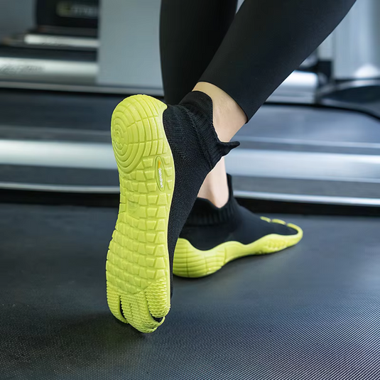 Schwarze Barfußschuhe mit gelber Sohle auf Laufband, minimalistisches Design, ideal für Fitness und Laufen, flexible und rutschfeste Sohle.