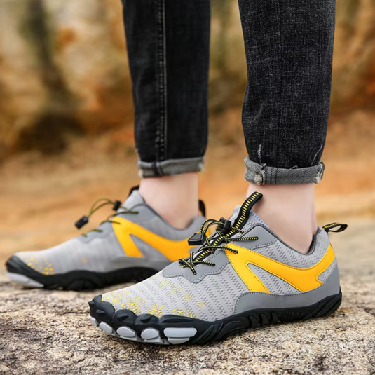 Graue und gelbe Barfußschuhe auf einem Felsen, getragen von einer Person in hochgekrempelten Jeans. Outdoor-Sportschuhe, minimalistisch, flexibel.