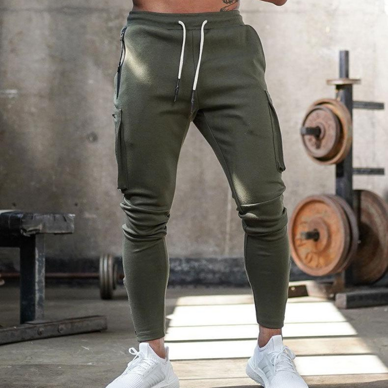Laden Sie das Bild in Galerie -Viewer, Grüne Jogginghose im Fitnessstudio, kombiniert mit weißen Turnschuhen. Hantelbank und Gewichte im Hintergrund. Sportmode, Fitness, Herrenbekleidung.
