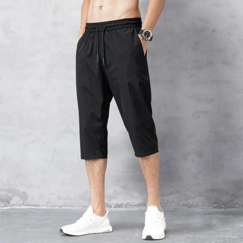 Laden Sie das Bild in Galerie -Viewer, Mann trägt schwarze Dreiviertelhose und weiße Sportschuhe vor grauem Hintergrund. Sportliche Herrenmode, bequeme Freizeitkleidung, Fitness-Outfit.
