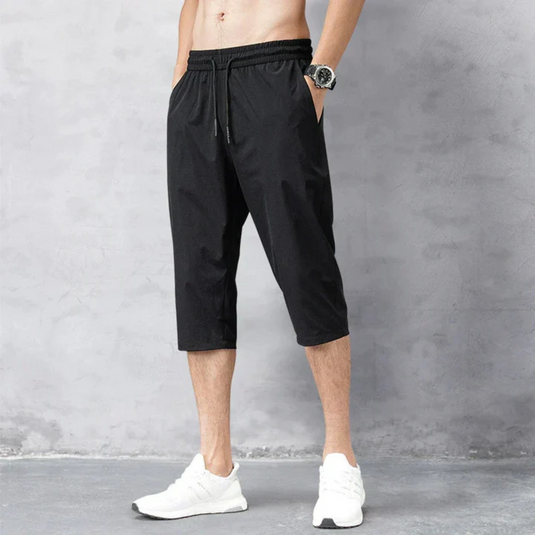 Mann trägt schwarze Dreiviertelhose und weiße Sportschuhe vor grauem Hintergrund. Sportliche Herrenmode, bequeme Freizeitkleidung, Fitness-Outfit.