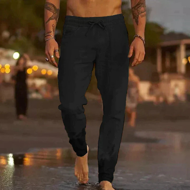 Laden Sie das Bild in Galerie -Viewer, Mann in schwarzen Jogginghosen am Strand bei Sonnenuntergang, barfuß im Sand. Freizeitmode, Strandspaziergang, entspannte Atmosphäre.

